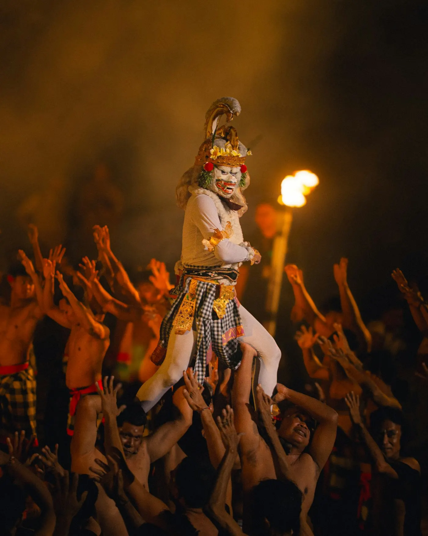 Kecak at night, a masked figure lifted above a circle of chanting men by torchlight